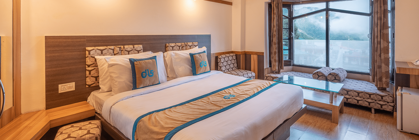 A hotel room with a large bed, a window overlooking a mountain view, and a seating area - DLS Meghavan Resort